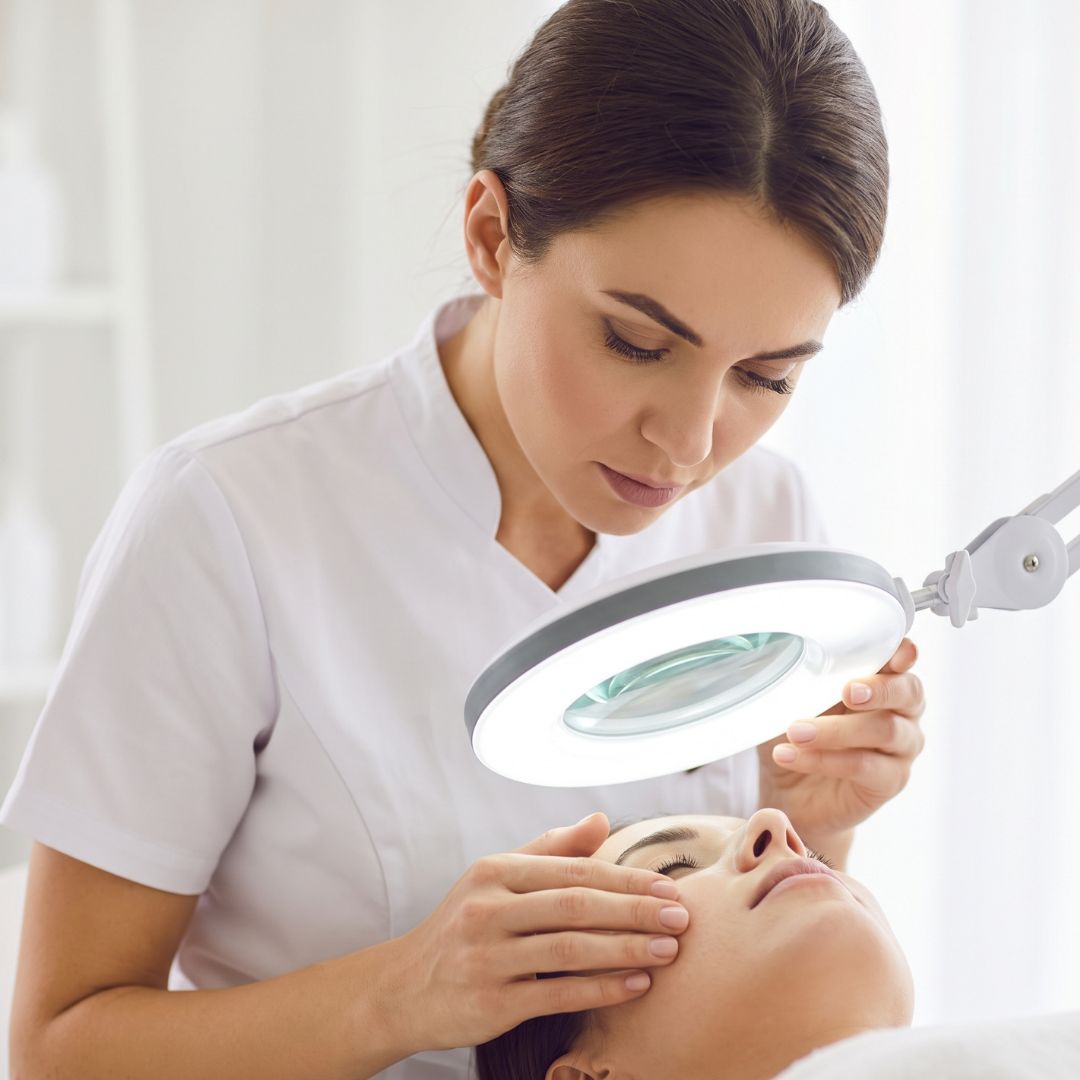 An esthetician uses a magnifying lamp to examine a client's skin.