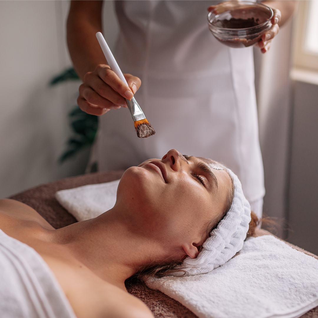 A gloved hand uses a brush to apply a detailed product treatment during a facial procedure.